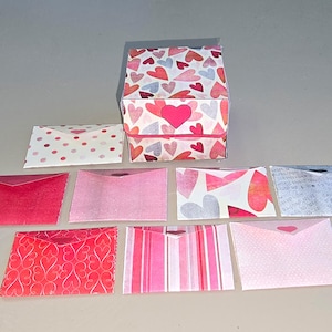 Könnte beinhalten: Eine kleine, quadratische Geschenkbox und mehrere Umschläge, alle mit Herzmustern in Rot-, Rosa- und Weißtönen verziert. Die Box hat einen großen Herz-Ausschnitt auf der Vorderseite. Die Umschläge variieren im Design.