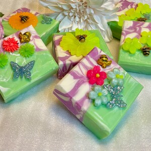 May include: Square handmade soaps with green and white swirls, decorated with colourful flowers, butterflies, and bee embellishments. The soaps are arranged on a white surface, with pink roses and white flowers in the background.