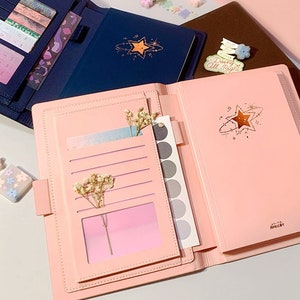Puede incluir: Dos diarios de cuero rosa y azul abiertos con un diseño de estrella y planeta. Los diarios tienen bolsillos y una ventana de plástico transparente para una foto o una nota. Los diarios están sobre una superficie rosa con flores y pegatinas decorativas.