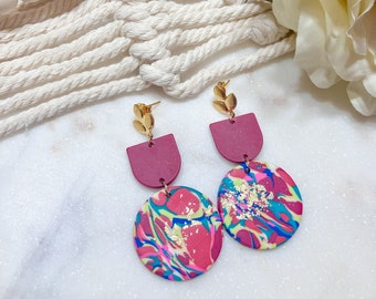 Puede incluir: Pendientes colgantes con detalles de hojas doradas. Los pendientes presentan una forma de semicírculo granate, conectada a un colgante circular con un colorido patrón de remolino de rosa, azul, amarillo y motas doradas.