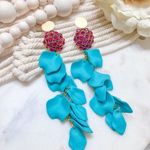 Może przedstawiać: Para turkusowych kolczyków z płatkami z złotymi akcentami. Kolczyki mają złotą tarczę i skupisko różowych kryształków na górze. Kolczyki są umieszczone na białym i beżowym tle.