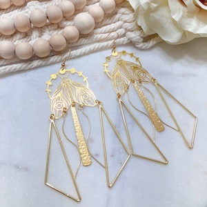 Puede incluir: Pendientes de tono dorado con un diseño geométrico que presenta un motivo de mariposa y triángulos colgantes.