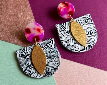 May include: A pair of contemporary earrings. Each earring has a pink and white marbled disc, a gold leaf-shaped charm, and a black and white patterned semi-circle. The earrings are displayed against a colourful background.