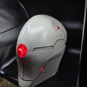 May include: A white helmet with black lines and red lights. The helmet is made of a smooth, hard material and has a rounded shape.