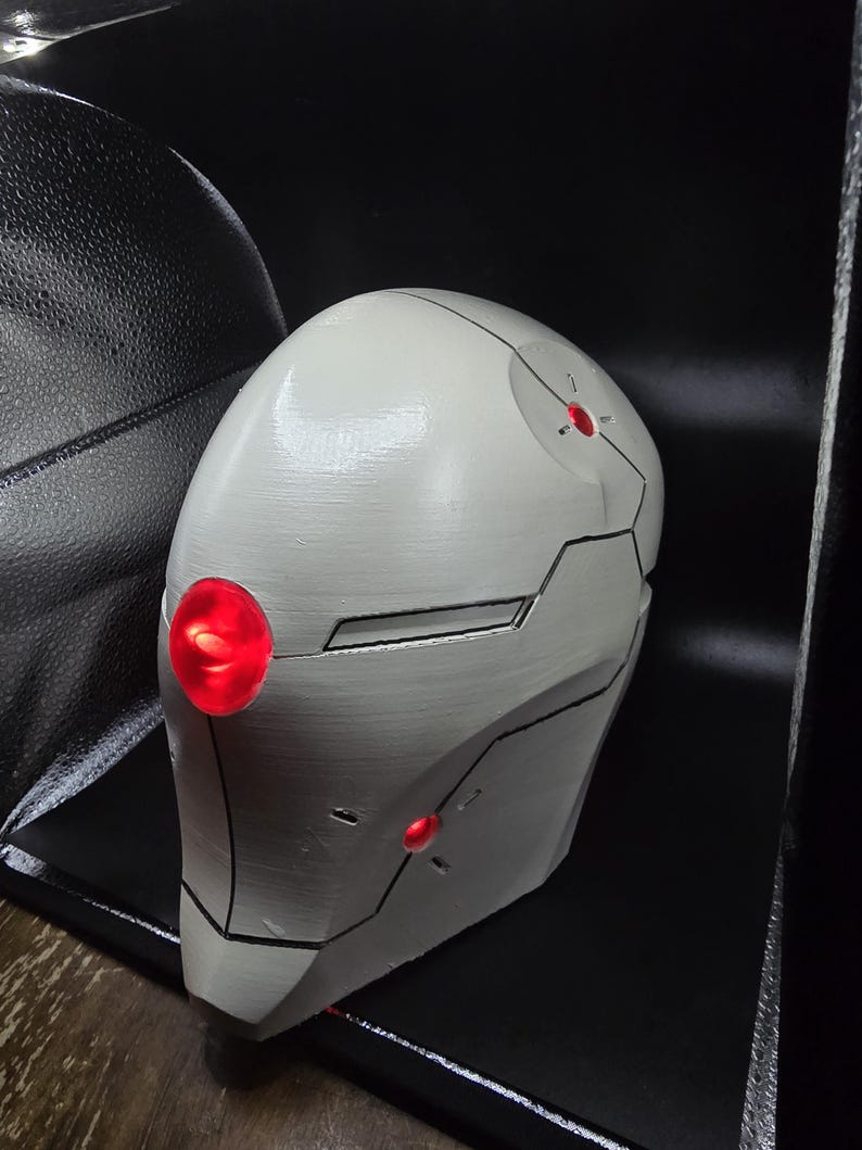 May include: A white helmet with black lines and red lights. The helmet is made of a smooth, hard material and has a rounded shape.