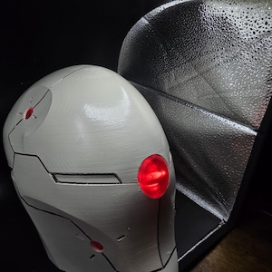 May include: A white helmet with black lines and red lights. The helmet is made of a smooth, hard material and has a rounded shape.