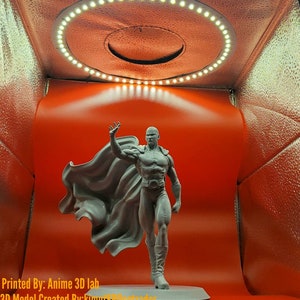 May include: A 3D printed grey figurine of a superhero with a cape, standing on a grey base. The figurine is in a white box with a ring of white lights around the top. The text "Printed By: Anime 3D lab" and "3D Model Created By: kimeto888gtrader" is visible at the bottom of the image.