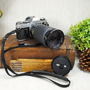 May include: A silver and black Canon AE-1 35mm film camera with a black lens, a black camera strap, and a circular black lens cap labeled Pro Master. A yellow folding wooden ruler is visible, showing measurements in inches up to 6 inches.