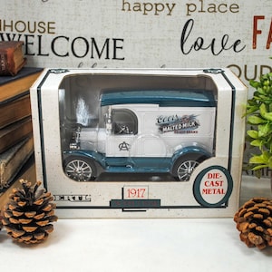 May include: A die-cast metal model of a vintage Coors Malted Milk delivery truck, white with teal accents, displayed in its original box. The box features the year 1917. The image includes pinecones and a stack of antique books.