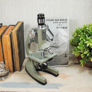 May include: A sage green Tasco zoom microscope with a silver eyepiece and objective lenses. The microscope is accompanied by its original instruction sheet featuring the text "EXPLORE NEW WORLDS within our world with a tasco" and "ZOOM MICROSCOPE KIT".