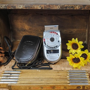 May include: A vintage light meter with a black leather case and a wooden folding ruler are displayed. The light meter is silver with a circular dial. Three yellow sunflowers with brown centers add a pop of color.