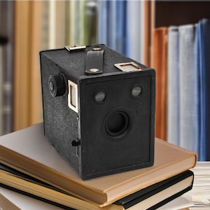 May include: A vintage black box camera with a leather handle and a lens, resting on a stack of books. The camera has a circular lens and two smaller circular openings. The books have tan and black covers.