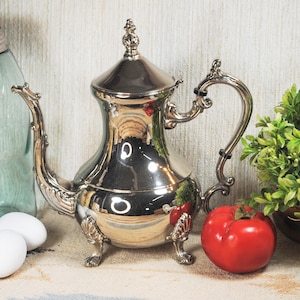 May include: A polished silver-tone metal teapot featuring an ornate curved handle, a decorative spout, and a lid topped with a finial. The teapot stands on three detailed feet and has a reflective surface that shows the surrounding environment.