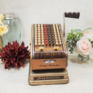 May include: Vintage Paymaster cash register with a brown wooden finish, featuring a handle and numerous number keys. The register has the words "Locked Protection" and "Paymaster" on it. The image also includes flowers in vases.