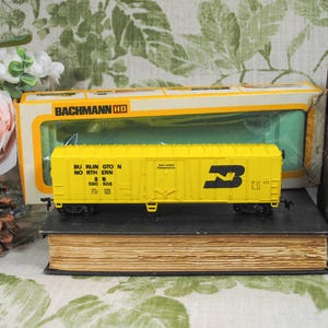May include: A yellow Burlington Northern HO scale model train car with black lettering and a logo. The train car is displayed on a vintage book, with a Bachmann box in the background. A pinecone and flowers are also in the image.