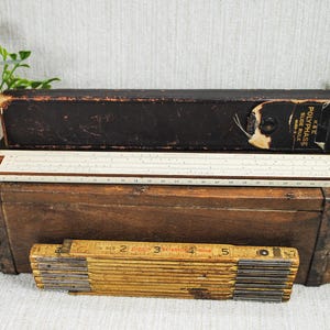 May include: A beige K&E Polyphase slide rule with a black storage case, positioned above a yellow folding wooden ruler with metal hinges. The slide rule features numerical scales and markings, while the folding ruler displays imperial measurements.