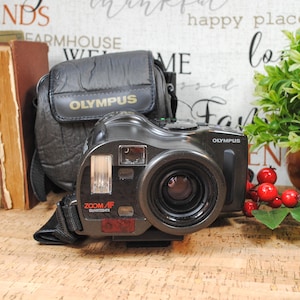 May include: A black Olympus Zoom AF Quartzdate camera with a lens, flash, and strap. The camera is next to a black case with the Olympus logo in gold. A small plant and books are in the background.