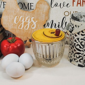 May include: A rustic farmhouse-themed still life featuring a yellow-topped butter churn, a red tomato, and three white eggs. A decorative chicken-shaped sign with the word "eggs" is visible. A black and white decorative bucket is also present.