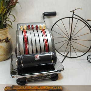 May include: Vintage cash register with silver and red details, featuring a black handle and a display of numbers. The Hedman Company logo is visible. A decorative penny-farthing bicycle and a ceramic vase are in the background.