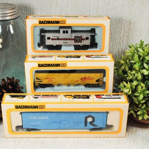 May include: Three Bachmann HO scale model train cars in their original boxes. The boxes are white and orange with clear windows. The train cars are a mix of colors, including blue, yellow, and silver.