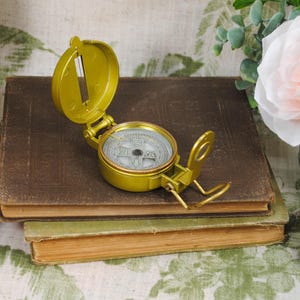 May include: A vintage brass compass with an open lid, resting on a stack of old books. The compass face is visible, and the books have aged, textured covers. A decorative floral arrangement and pinecone are in the background.