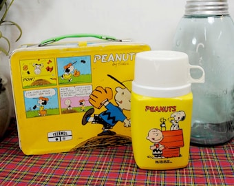 May include: A yellow vintage Peanuts lunchbox and thermos set. The lunchbox features comic panels and the word "Peanuts" by Schulz. The thermos has a white lid and handle, with Peanuts characters. The set is on a red and black plaid surface.