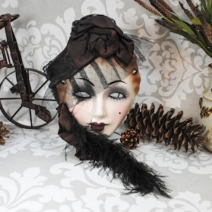 May include: A porcelain mask with a black and white striped veil, brown hat, and black feather. The mask has painted white eyes, a red mouth, and a light pink blush on the cheeks.