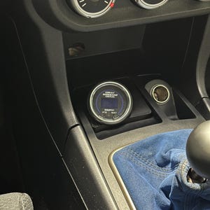 May include: A car's interior featuring a Cobalt Wideband Air/Fuel gauge installed in the center console. The gauge has a blue and white display. The console is black, with a blue fabric gear shift boot visible.