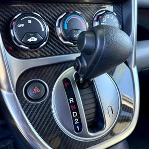 May include: Close-up of a car's interior, showcasing the gear shift lever, climate control dials, and hazard light button. The gear shift is black, with a silver surround and the letters P, R, N, D, 2, and 1. The dashboard has a carbon fiber pattern.