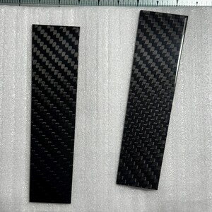 May include: Two rectangular carbon fiber strips with a distinct black and grey woven pattern. The strips are positioned on a white foam sheet next to a ruler showing measurements in inches, with the left strip measuring approximately 4 inches long.