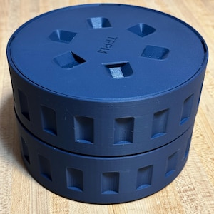 May include: A two-piece, navy blue, cylindrical container. The top piece has eight rectangular cutouts and the letters "TFPM". The bottom piece has a series of rectangular cutouts around the sides. The container is sitting on a wooden surface.