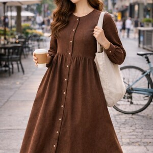Casual button-down gathered dress sewing pattern shown on model in realistic lifestyle setting, featuring a relaxed bodice, full button placket, long sleeves, and soft gathered skirt. This beginner friendly women’s PDF sewing pattern is designed for cotton poplin, linen, and other light woven fabrics for comfortable daily wear.