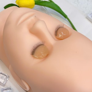 Pode incluir: Uma cabeça de manequim de treino com cílios postiços aplicados. O manequim é de cor pêssego claro, com os olhos fechados. Os cílios postiços são pretos e estão presos a uma substância clara semelhante a gel. Uma flor amarela e folhas verdes ao fundo.
