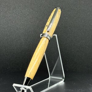 May include: A wooden pen with a light brown and black grain pattern, accented with silver-colored metal hardware. The pen is displayed on a clear acrylic stand against a dark background. The pen's tip is black.