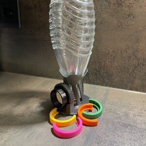 Könnte beinhalten: Ein klarer Wasserflaschenspender aus Kunststoff mit grauem Sockel und schwarzer Düse. Der Spender hat eine Reihe farbiger, gebogener Ringe in Pink, Gelb, Orange und Grün. Der Spender ist für eine Stapel von Wasserflaschen konzipiert.