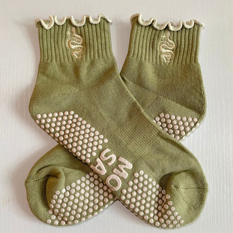 May include: A pair of olive green ankle socks with a cream-colored, scalloped trim. Each sock features a beige embroidered snake design and white non-slip grips on the soles. The word "MOSA" is printed on the sole.