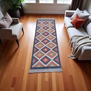 Puede incluir: Una alfombra de pasillo rectangular con un patrón geométrico de diamantes en tonos rojos, azules, amarillos y blancos. La alfombra tiene un borde azul oscuro y flecos en ambos extremos. La alfombra está colocada sobre un suelo de madera.