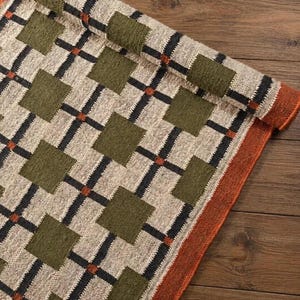 May include: A woven textile runner featuring a grid pattern of black lines and small orange squares on a cream background, interspersed with larger olive green squares. The runner has a solid orange border along the edge and is partially rolled up.