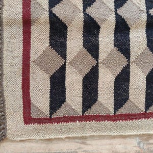 May include: Close-up of a woven rug with a geometric pattern. The rug features a repeating design of three-dimensional shapes in black, beige, and gray. A thin red border frames the design, and the edge has a fringe.