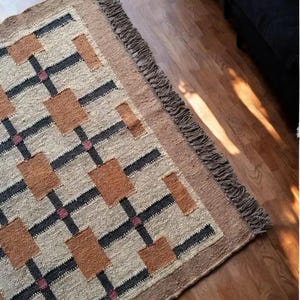 Op de afbeelding: Een rechthoekig vloerkleed met een geometrisch patroon. Het kleed heeft een beige achtergrond met bruine, zwarte en roze accenten. De rand en franjes zijn bruin. Het kleed ligt op een houten vloer.