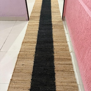 Puede incluir: Una alfombra larga y rectangular de yute con una franja negra central. La alfombra tiene un color beige natural a ambos lados de la franja negra. La alfombra está colocada sobre un suelo de baldosas blancas.