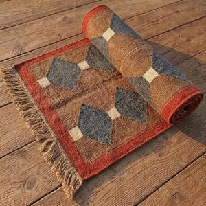 Peut inclure: Un tapis tissé rectangulaire présentant un motif géométrique de losanges gris foncé et marron séparés par de petits carrés blancs. Le tapis possède une bordure épaisse orange brûlé et une extrémité à franges, partiellement enroulé.