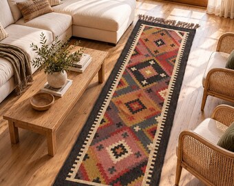 May include: A long rectangular runner rug featuring a geometric pattern with diamonds and crosses in shades of red, pink, yellow, and dark gray. The rug has a cream-colored border and is finished with knotted fringe tassels on both ends.