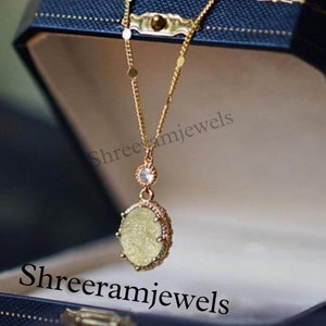 Puede incluir: Un collar de cadena dorada con un colgante redondo que presenta una piedra verde claro brillante rodeada de piedras más pequeñas. El colgante está colgando en una caja de joyería de terciopelo azul con el texto "Shreeramjewels" impreso en la caja.