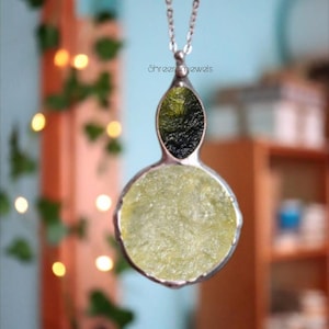 Puede incluir: Un collar con colgante de plata con una piedra preciosa redonda verde claro en la parte inferior y una piedra preciosa ovalada verde oscuro en la parte superior. El collar cuelga de una cadena de plata. El texto "Shreer Jewels" es visible.