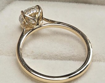 May include: A gold engagement ring with a large, round, clear gemstone. The ring has a thin band and a four-prong setting. The ring is set in a white velvet ring box.