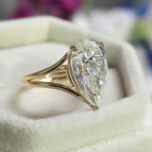 Op de afbeelding: Een verlovingsring met een peervormige diamant, gezet in een gouden band met een gesplitste schouder. De ring ligt in een witte fluwelen ringdoos.