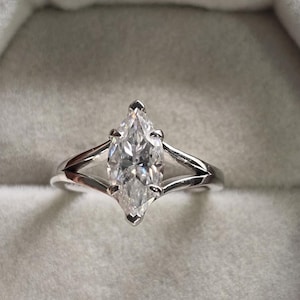 May include: A marquise cut diamond engagement ring set in a white gold band with a split shank design. The ring is displayed in a white velvet ring box.