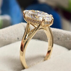 Puede incluir: Un anillo de oro con una gran piedra preciosa transparente de talla marquesa en un engaste de garras. La banda es de oro amarillo pulido y la cesta que sostiene la piedra central está decorada con una hilera de pequeñas piedras redondas.