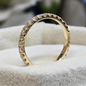 Könnte beinhalten: Ein Goldband mit einer Reihe kleiner Diamanten, die in das Metall gefasst sind. Das Band ist in einer weißen Schachtel abgebildet.
